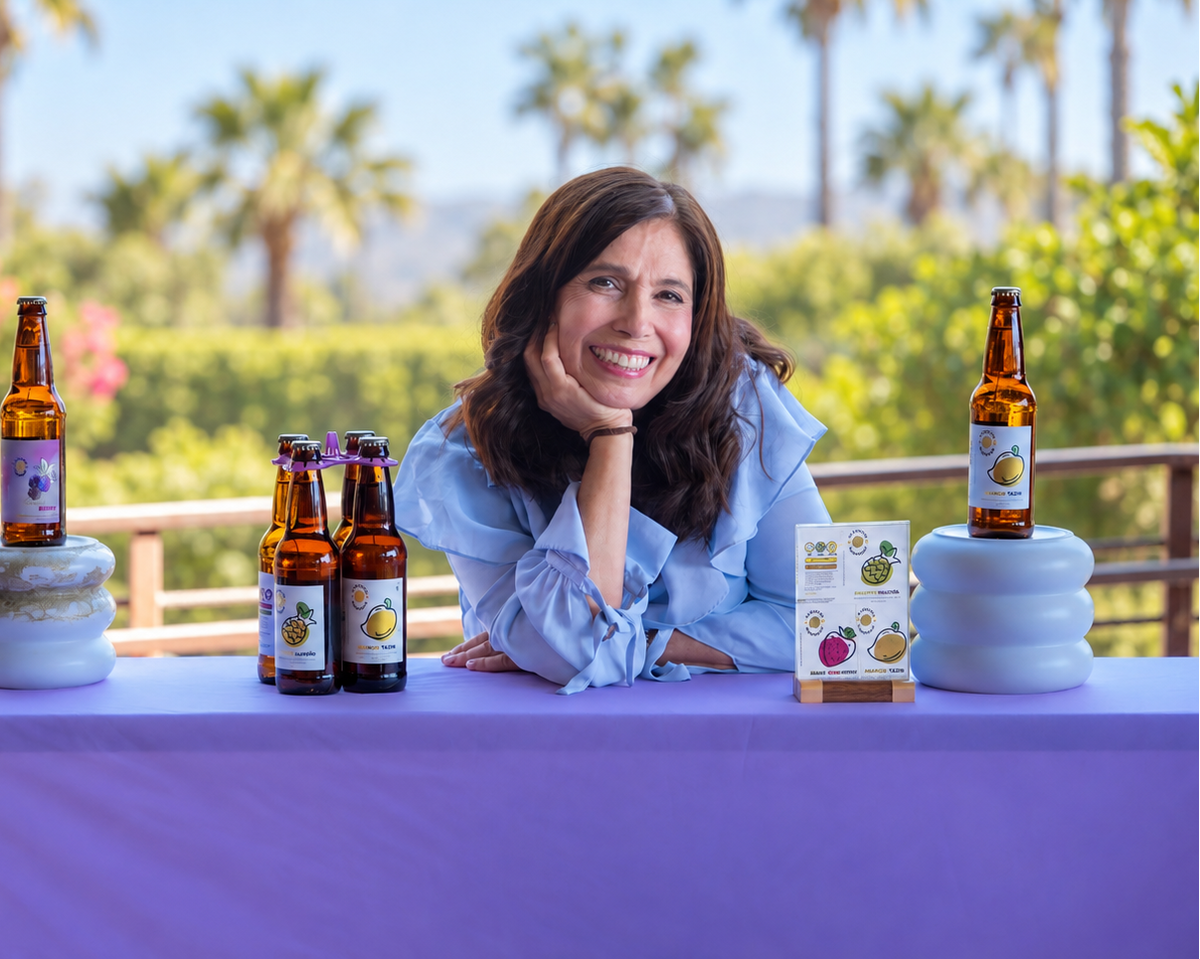 A smiling woman in a light blue shirt leans on a lavender tablecloth-covered table displaying multiple bottles of Fruit of the Loom Gummy Bears, set against a sunny outdoor background with palm trees and a railing.